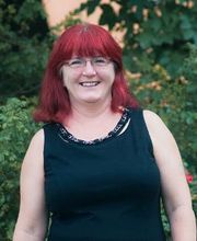 Frau mit rotem Haar und Brille lächelt freundlich. Sie steht vor grünen Pflanzen.