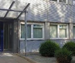 Ein modernes Gebäude mit Metallfassade. Links befindet sich ein Eingang mit blauem Türrahmen und Glasüberdachung. Rechts steht ein Schild mit Logos und „WBS Training“. Vor den Fenstern sind Büsche und ein Kiesbett angelegt. Der Bereich wirkt gepflegt.