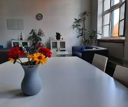 Aufenthaltsraum mit Sesseln, Sofa, Zimmerpflanzen sowie einem Tisch auf dem eine Vase mit bunten Blumen steht.