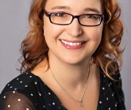 Portrait einer Mitarbeiterin mit hellbraunen, gewellten Haaren und Brille, die freundlich in die Kamera lächelt.