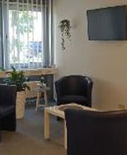  Ein gemütlicher Wartebereich mit vier schwarzen Sesseln und einem weißen Couchtisch. An der Wand hängt ein Fernseher, und Pflanzen dekorieren den Raum. Große Fenster mit Jalousien lassen Licht hinein. Der Boden ist mit grauem Teppich ausgelegt.