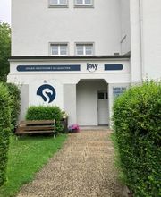Steinweg zum weißen Gebäude, vorbei an grünem Rasen und einer Sitzbank.