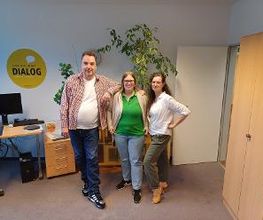 Ein Mann und zwei Frauen stehen nebeneinander und posieren für ein Foto. Neben ihnen befindet sich ein Schreibtisch mit Computerarbeitsplatz.