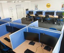 Ein Schulungsraum mit mehreren Arbeitsbereichen, die durch blaue Trennwände voneinander getrennt sind. Jeder Arbeitsplatz besteht aus 2 PC-Bildschirmen, Tastatur und Maus sowie Headset. Blaue Trennwände.