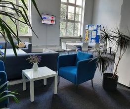 Aufenthaltsraum mit blauen Sesseln, grauen Sofas, einem Tisch mit 7 Stühlen, Fernseher an der Wand sowie großen Fenstern, Zimmerpflanzen und einer Moderationstafel.