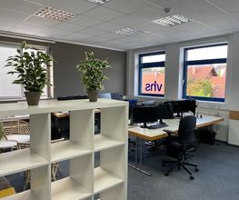 Ein Schulungsraum mit mehreren Arbeitsbereichen, die jeweils mit 2 PC-Bildschirmen, Tastatur und Maus sowie Headset ausgestattet sind. Im Vordergrund steht ein weißes Regal mit Topfpflanzen.