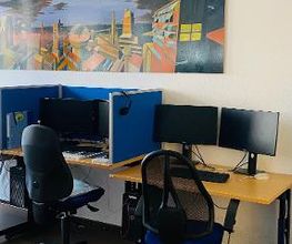 Arbeitsplatz mit zwei Bildschirmen, Maus, Tastatur, Headset. Darüber ein buntes Bild mit Gebäuden.