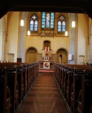 Unter der Empore angekommen öffnet sich der Blick auf den Kirchenraum, den Altar und den flachen Chorabschluss mit dem großen Drillingsfenster, dessen Originalverglasung aus der Bauzeit vom September 1899 bis Oktober 1900 erhalten geblieben ist. Auch die Kirchenbänke stammen noch aus dieser Zeit. Das Lilienmotiv aus den Terrakotta- Fußbodenfliesen wiederholt sich mehrfach in der Kirche.
