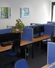 Ein Schulungsraum mit mehreren Lernplätzen, die durch blaue Trennwände voneinander getrennt sind. Jeder Arbeitsplatz besteht aus 2 PC-Bildschirmen, Tastatur und Maus sowie Headset.