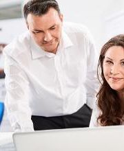 Dame, weiß gekleidet, dunkle lange Haare, sitzt am Tisch mit einem Notebook und lächelt ins Bild. Neben ihr steht ein Herr, dunkle Haare, weißes Hemd, und zeigt etwas am Notebook.