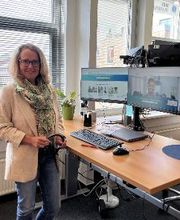 Eine Frau mit Brille und hellbeigem Blazer steht an einem höhenverstellbaren Schreibtisch. Auf dem Tisch stehen ein Monitor, Tastatur, Maus und eine Karaffe. Der Monitor zeigt eine Videokonferenz. Im Hintergrund sind Fenster mit Jalousien.