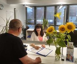 2 Kolleg:innen unterhalten sich über Formulare. Auf dem Tisch steht eine Vase mit Sonnenblumen.
