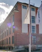 Außenansicht vom Schulungsgebäude an einem hellen Tag. Die Fassade ist rot und weiß geklinkert. Im Vordergrund sind 2 Fahnenmäste mit der Aufschrift „WBS TRAINING“ aufgestellt.