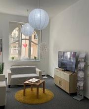 Aufenthaltsraum mit 2 Sofas und einem Beistelltisch. Auf der rechten Seite ein Regal mit TV und einem Flyerständer. Von oben hängen 2 runde Lampen herunter. Eine Stehlampe befindet sich neben einem Sofa.