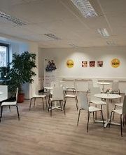 Aufenthaltsbereich mit 4 Tischen und jeweils 4 Stühlen und links und rechts sind große Bodenpflanzen