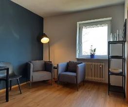 Aufenthaltsraum mit 2 Sesseln vor einer Stehlampe, daneben einem Regal mit Deko und einem weiteren Tisch mit Stuhl