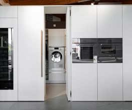 Moderne, grifflose Hochschrankküche mit integrierter Geheimtür zu einem praktischen Hauswirtschaftsraum. Hinter der flächenbündigen Schrankfront verbergen sich Waschmaschine, Stauraum und Arbeitsbereich – eine elegante Lösung für ein aufgeräumtes, funktionales Wohnkonzept.