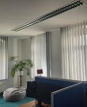 Schulungsraum mit 3 Fenstern und Vorhängen. Eine Pflanze steht vor einem Sitzsack. Rechts sind mehrere Arbeitsplätze erkennbar.