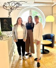 Ein Teambild. 3 Frauen stehen im hellen Aufenthaltsraum vor dem runden Fenster. Im Hintergrund ist ein kunstvolles Bild eines Leuchtturms zu sehen.