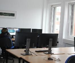 Schulungsraum mit Schreibtischen auf denen sich jeweils zwei Bildschirme, eine Tastatur, Maus und Headset befinden. In dem Raum befindet sich eine Person, die lernt. Fensterfront.