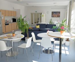 Helle, voll ausgestattete Teilnehmerküche mit Getränkeautomaten. 2 blaue Sofas und weiße Tische mit Stühlen. Mehrere Zimmerpflanzen und ein Bild an der Wand.