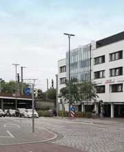 Eine Straßenecke mit einem weißen mehrstöckigen Gebäude, das Büros und Geschäfte beherbergt, darunter „ver.di“. Links ist eine Unterführung zu sehen, vor der Autos warten. Es gibt Ampeln, Verkehrsschilder und einen Fahrradweg. Der Himmel ist bewölkt.