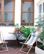 Eine kleine, gemütliche Terrasse mit Tisch, Holzliegestühlen und Sonnenschirm sowie einigen, grünen Pflanzen.