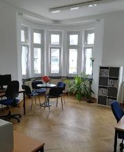 Heller Raum mit einigen Arbeitsplätzen sowie einer Sitzgelegenheit bestehend aus einem runden Tisch, auf dem Blumen stehen, und 4 Stühlen.