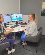 Eine Beratungssituation. Zwei Frauen sitzen an einem Tisch vor 2 Bildschirmen und unterhalten sich. Hellgraue Wand mit Textildekoration.