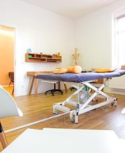 Ein geräumiger Behandlungsraum mit einer zentral platzierten, höhenverstellbaren Therapieliege mit dunkelgrauem Bezug. Mehrere ergonomische, orangefarbene Polsterkissen liegen darauf. Der Raum hat helle Wände und einen warmen Holzfußboden. An der linken Wand hängt ein schwebendes Holzregal mit kleinen dekorativen Elementen. Direkt daneben steht ein großes anatomisches Skelettmodell. In der rechten Ecke befindet sich ein Bücherregal mit Fachliteratur und Pflegeprodukten. Große Fenster mit Jalousien sorgen für viel Tageslicht. Im Vordergrund stehen moderne weiße Stühle, die eine Sitzgelegenheit für Patienten oder Begleitpersonen bieten.