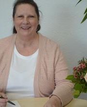 Eine Frau in rosa Strickjacke sitzt an einem Tisch und hält einen Stift über ein Notizbuch. Sie lächelt in die Kamera. Rechts auf dem Tisch steht ein Blumenstrauß in einer grünen Vase. Im Hintergrund sind eine weiße Wand und eine Pflanze zu sehen.