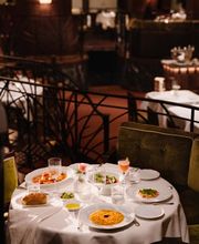 A white linen table laden with saffron risotto, pasta, cocktails, and appetizers, positioned next to a dark wrought-iron railing with green velvet seating.