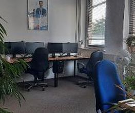 Schulungsraum mit 4 Arbeitsplätzen mit jeweils 2 Bildschirmen, einem Bürostuhl, Tastatur und Maus. Mehrere Pflanzen stehen im Raum, sowie ein Ventilator.