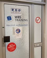 Glas-Eingangstür mit mehreren Aufklebern der WBS TRAINING wie einem Herzlich Willkommen Aufkleber und Hinweisen zur 100%igen Förderung.
