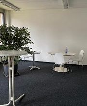 Heller Aufenthaltsraum mit weißen Tischen und Stühlen sowie einer großen, grünen Zimmerpflanze.