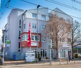 Dieses Bild zeigt das Gebäude auf der Carl-Benz-Straße von außen. Das Haus hat eine hellgraue Fassade und vor dem Gebäude stehen 2 Bäume sowie eine rote Fahne. 