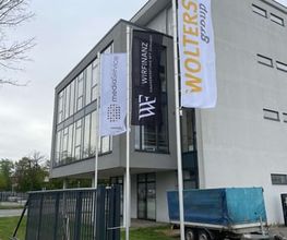 mediaService Bremen | Full-Stack Werbeagentur - Drei große weiße und schwarze Werbefahnen an Fahnenmasten wehen vor einem modernen Bürogebäude mit angeschlossenem Metalltor.