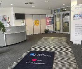 Eingangsbereich mit Empfang, Wartebereich, Werbeaufsteller und einer großen, grünen Zimmerpflanze. Auf dem Boden liegt ein Teppich mit der Aufschrift „WBS TRAINING – Herzlich Willkommen“