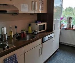 Eine kleine Küchenzeile mit beigen Schränken und dunkler Arbeitsplatte. Zu sehen sind ein Waschbecken, Herd, Mikrowelle und verschiedene Küchenutensilien. Rechts steht ein Wasserspender vor einem Fenster, durch das Bäume sichtbar sind.