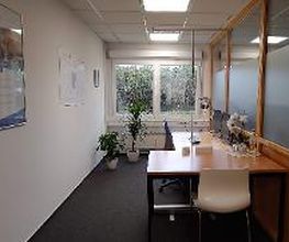 Ein schmaler Büroraum mit einem Schreibtisch, Stuhl und Computer. Auf dem Tisch stehen Dekorationen und eine Lampe. Links hängen Poster an der Wand, hinten sind Pflanzen vor einem Fenster. Rechts trennt eine Glastrennwand den Raum ab.