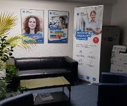 Aufenthaltsbereich mit Ledercouch und Sesseln, einer großen, grünen Zimmerpflanze sowie Druckstation. Zusätzlich steht ein Werbeaufsteller im Bereich.