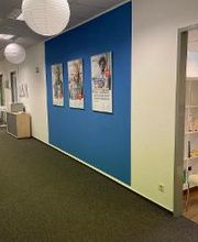 Flur mit einer blauen Wand an dem Plakate hängen. Blick in den gemütlichen Aufenthaltsraum mit Bücherregalen und Sitzgelegenheiten.