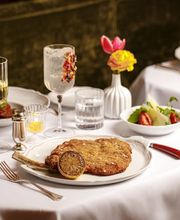 Classic Milanese-style veal cutlet with bone, breaded and golden, served with a grilled lemon half on a white plate, alongside a green salad, crusty bread, wine glasses, candlelight, and a small floral arrangement on a crisp white tablecloth.