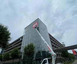 mediaService Bremen | Full-Stack Werbeagentur - Ein weißer Lkw mit einer ausgefahrenen Mateco-Arbeitsbühne montiert ein großes rotes Werbelogo an der Glasfassade eines modernen mehrstöckigen Gebäudes.