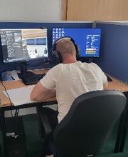 Mann sitzt am Arbeitsplatz mit 2 Bildschirmen und einer Tastatur. Er trägt ein Headset und vor ihm liegen mehrere Blätter. Rechts ein weiterer Arbeitsplatz.