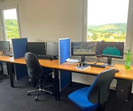 Drei Arbeitsbereiche vor Fenstern mit Blick ins Grüne. Die Arbeitsbereiche sind jeweils mit 2 Bildschirmen, Tastatur, Maus und Headset ausgestattet. Auf einem Tisch befindet sich ein Buch und eine grüne Wasserflasche.