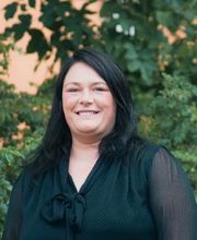 Portrait einer jungen Mitarbeiterin mit schwarzen Haaren, die draußen im Grünen steht.