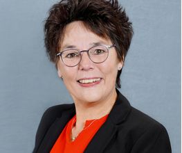 Eine Frau mit kurzen dunklen Haaren und Brille. Sie trägt einen schwarzen Blazer und eine rote Bluse.