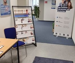 Eingangsbereich mit Teppich. Links ein Holztisch und ein blauer Stuhl, daneben ein Flyerständer. Auf der rechten Seite steht ein Roll Up Banner der WBS GRUPPE und im Hintergrund erkennt man ein Fenster sowie Zimmerpflanzen.
