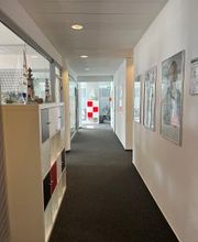 Ein heller Flur mit dunklem Teppichboden. Rechts hängen Poster an der weißen Wand, links stehen Regale mit Fächern. Am Ende des Flurs ist eine Glastür mit einer rot-weißen Markierung und ein Fluchtwegschild zu sehen.